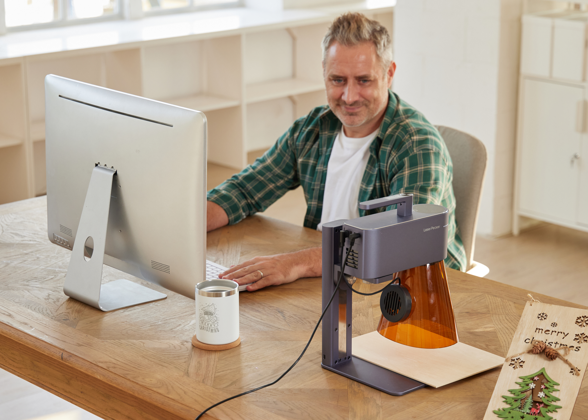 Kleinunternehmer entwirft individuelle Produkte am Computer, während der LaserPecker LP4 im Hintergrund Werkstücke graviert.