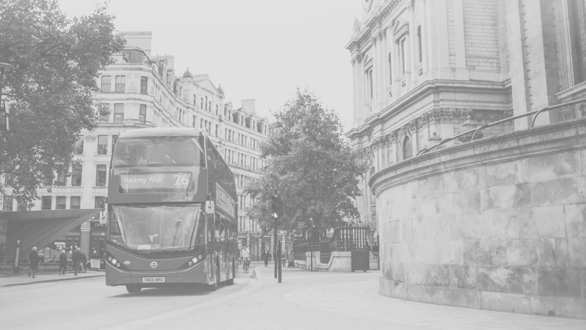 A street in the United Kingdom with a red bus driving along the road, flanked by historic white buildings and scattered pedestrians.