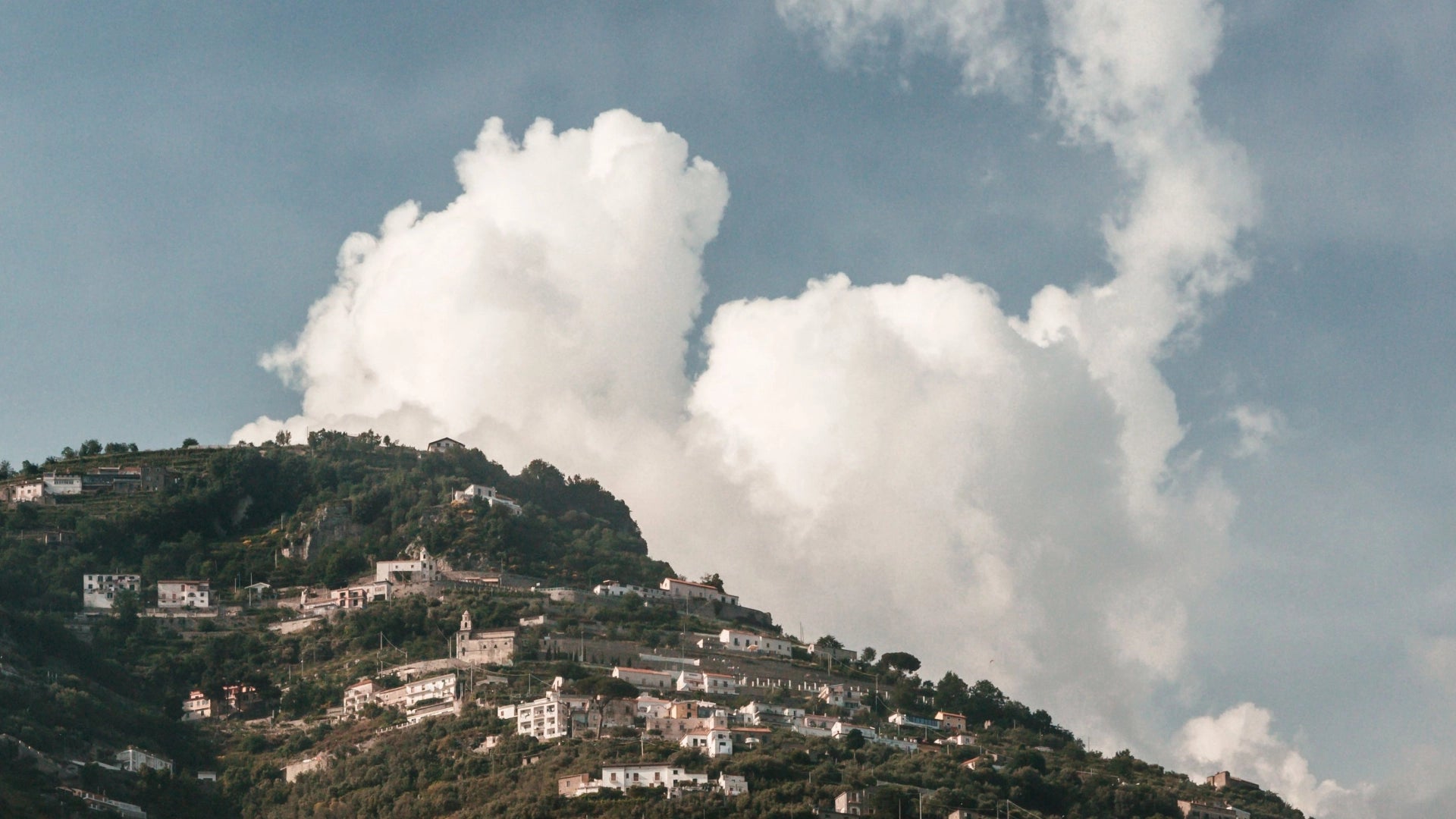 Coastal hills in Italy with several houses overlooking a vast sky filled with clouds.