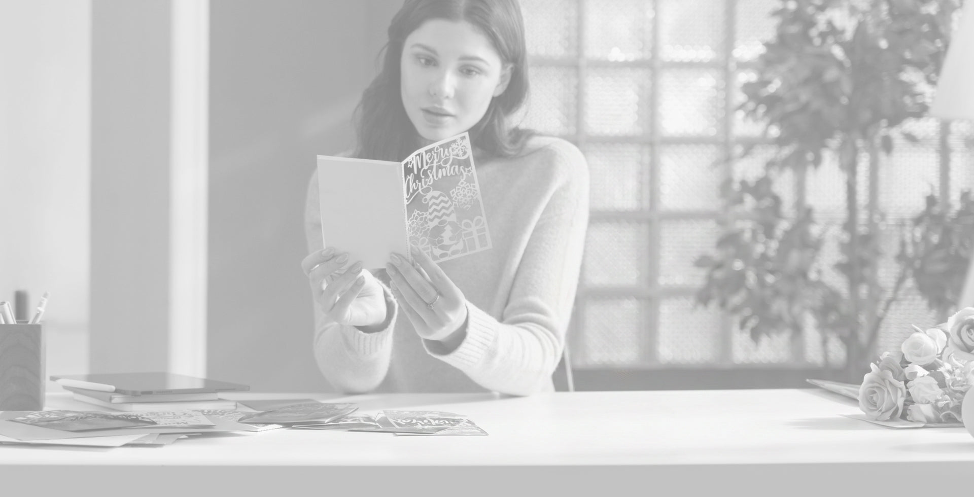 A woman holding a laser-cut hollow greeting card, inspecting the engraving results, with other engraved creative items and a laser engraving machine on the table.