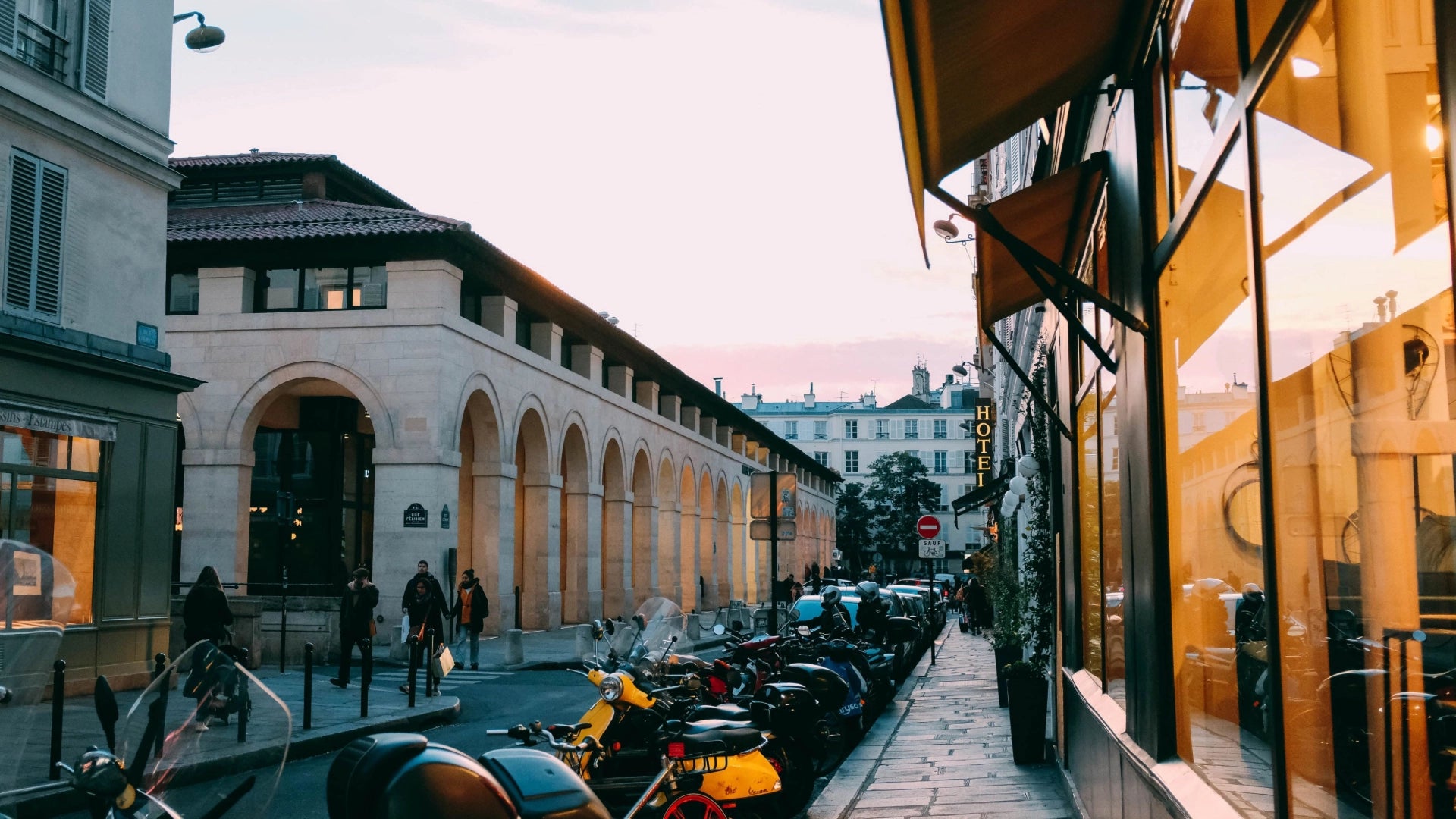 A street scene in France featuring architecturally striking buildings with arched entrances resembling a museum, with pedestrians walking and bicycles and scooters neatly parked along the roadside.